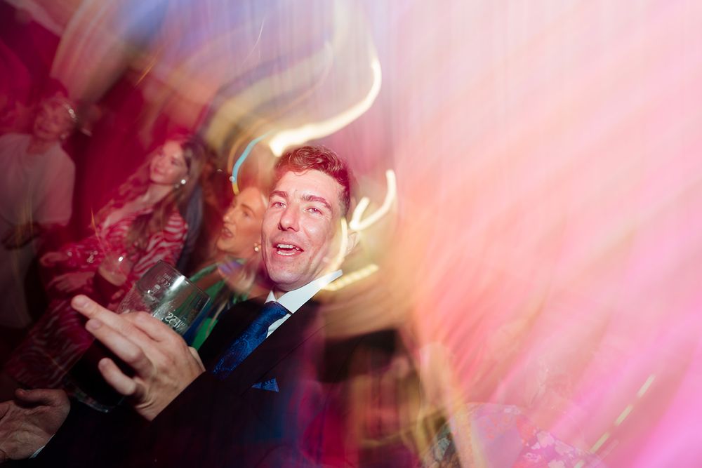 Wedding party guests dancing and celebrating with motion blur and pink lighting effects at a reception.