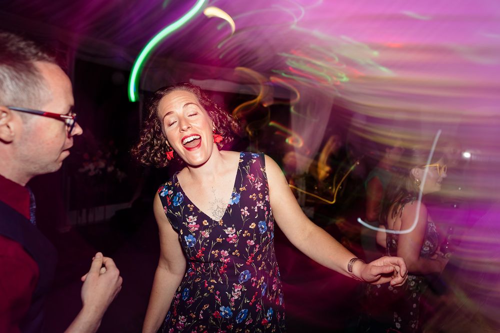 Person dancing enthusiastically in a floral dress under purple lighting at a party.
