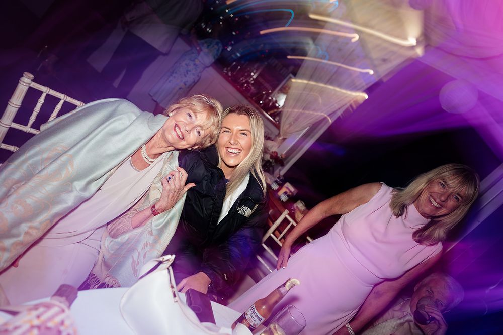 Wedding party celebrates on the dance floor with vibrant purple lighting and joyful expressions.