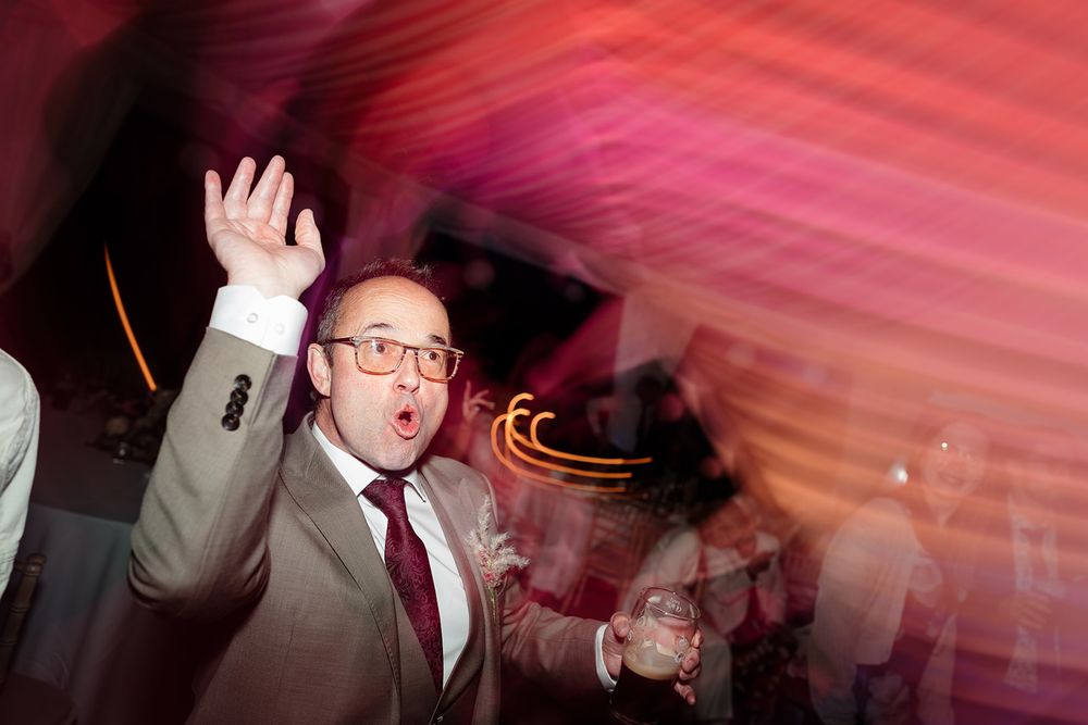 A person in glasses and suit dances enthusiastically at a party with colorful motion-blurred lighting in the background.