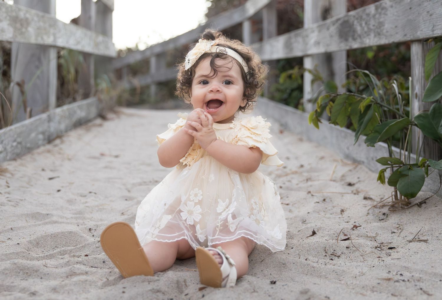 Adorable baby girl in cream lace dress sitting on sandy beach path smiling happily.