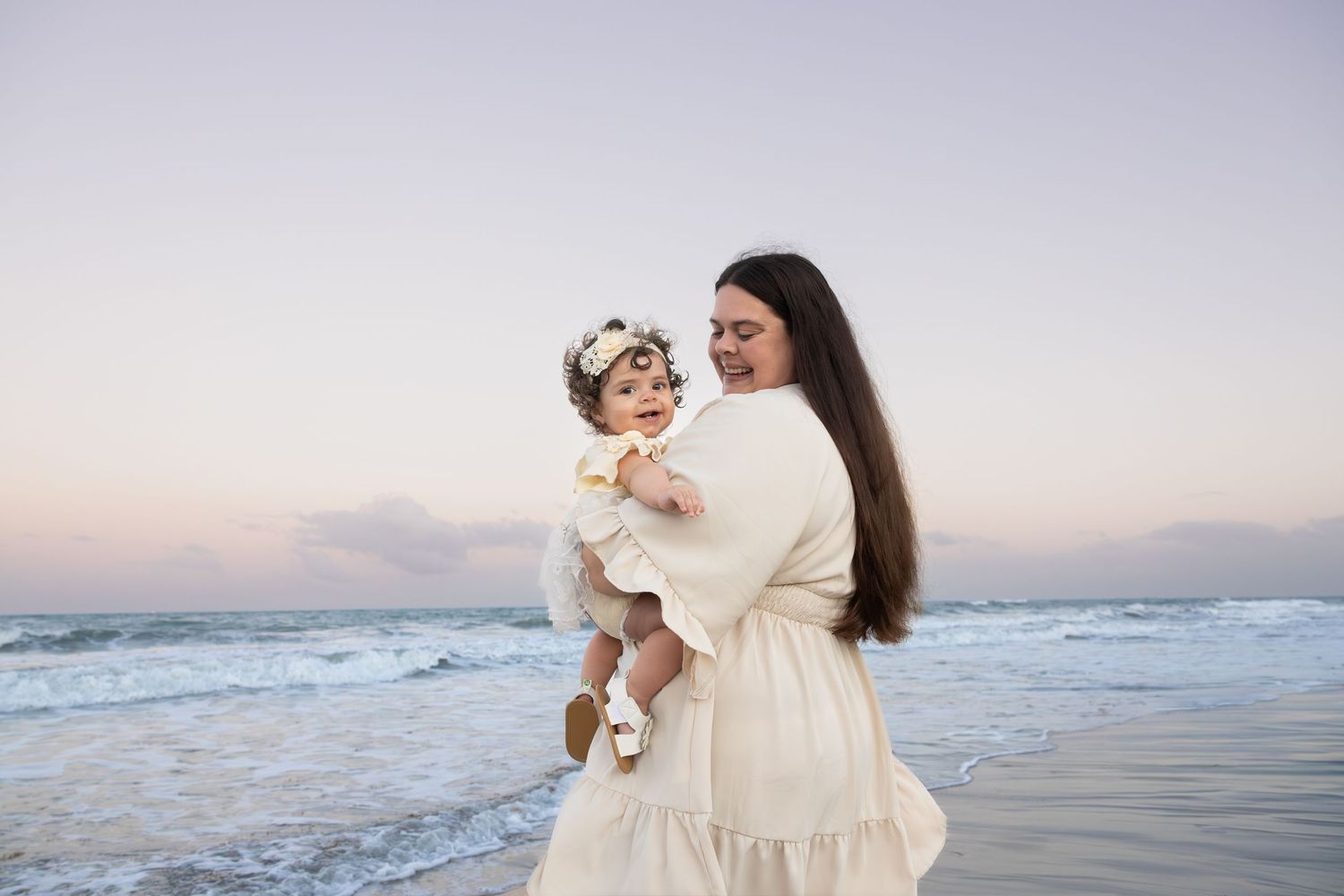Mother in cream dress holds baby with floral crown on beach at sunset.