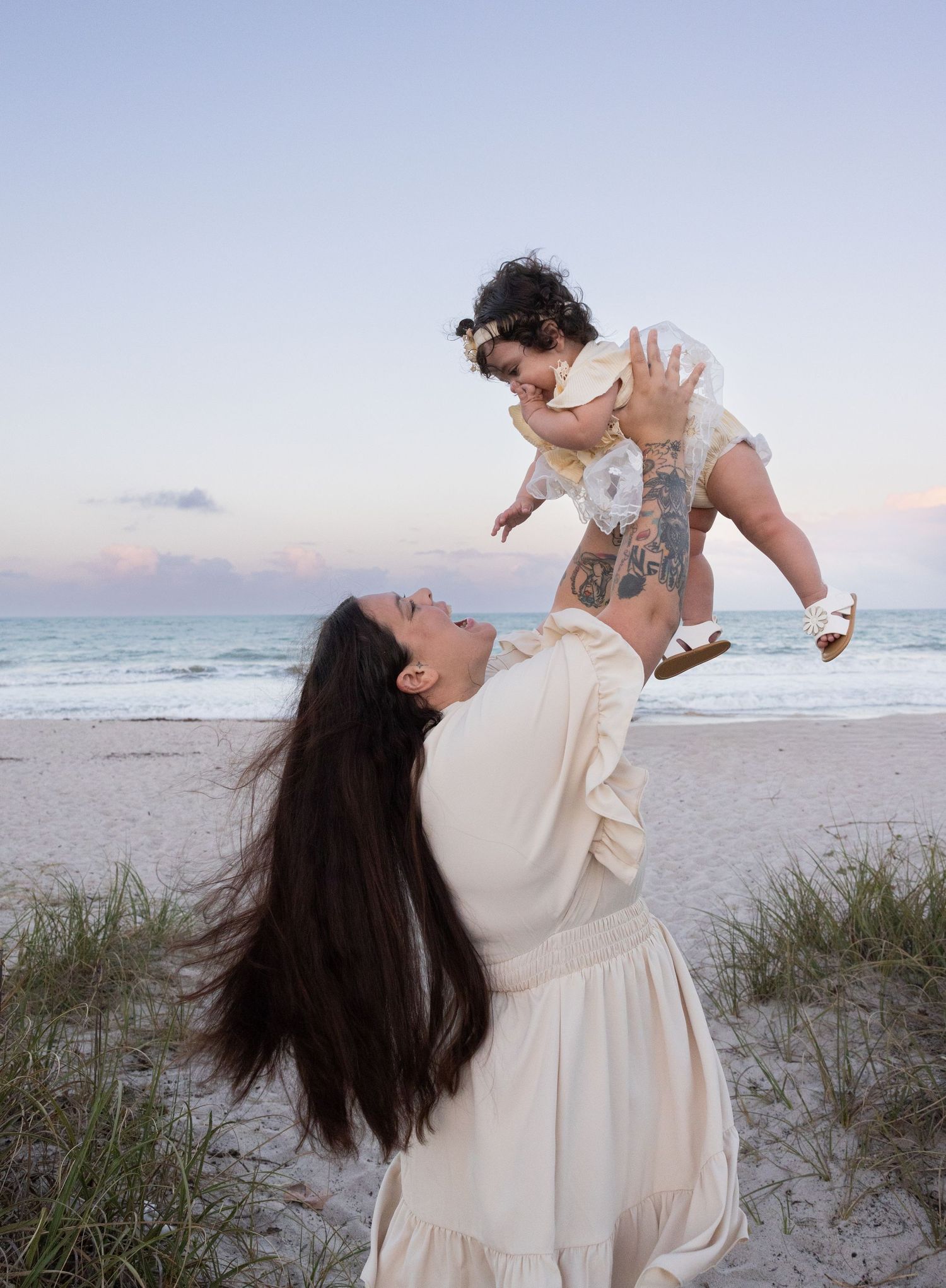 Mother lifting laughing baby in cream dress on a beach at dusk with ocean behind them.