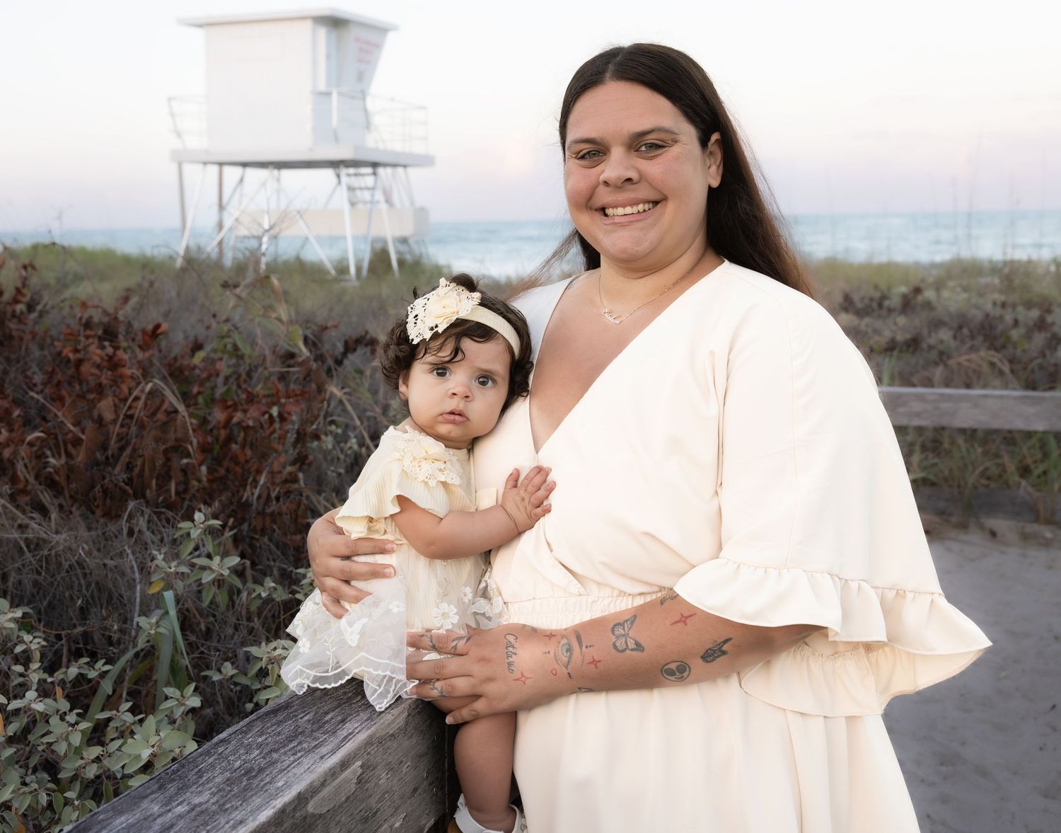 A mother in a cream dress holds her baby girl on a beach near a lifeguard tower at sunset.