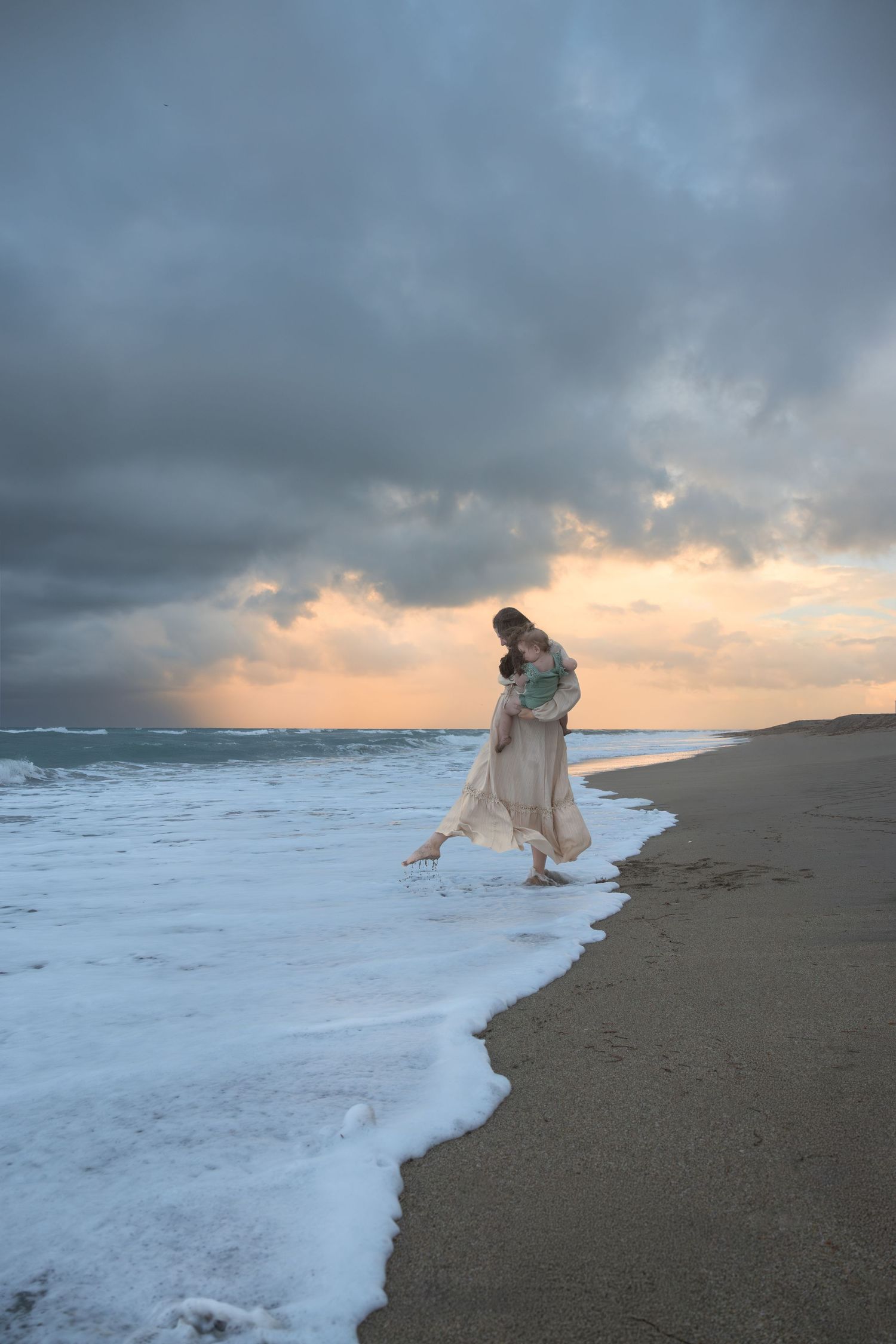 Woman in cream dress holds child while walking in ocean waves.