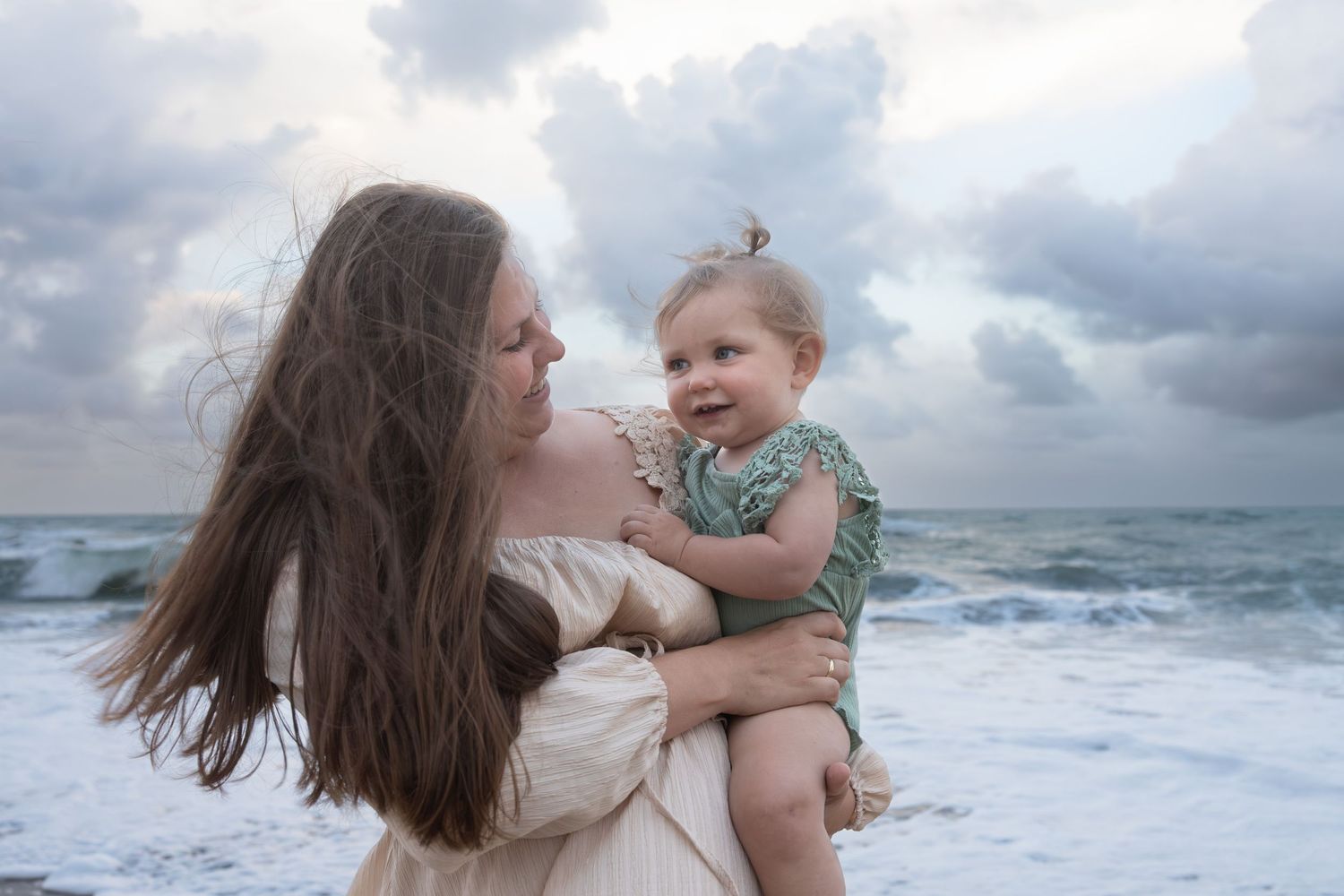 Mother with long hair holds smiling baby in green outfit at beach.