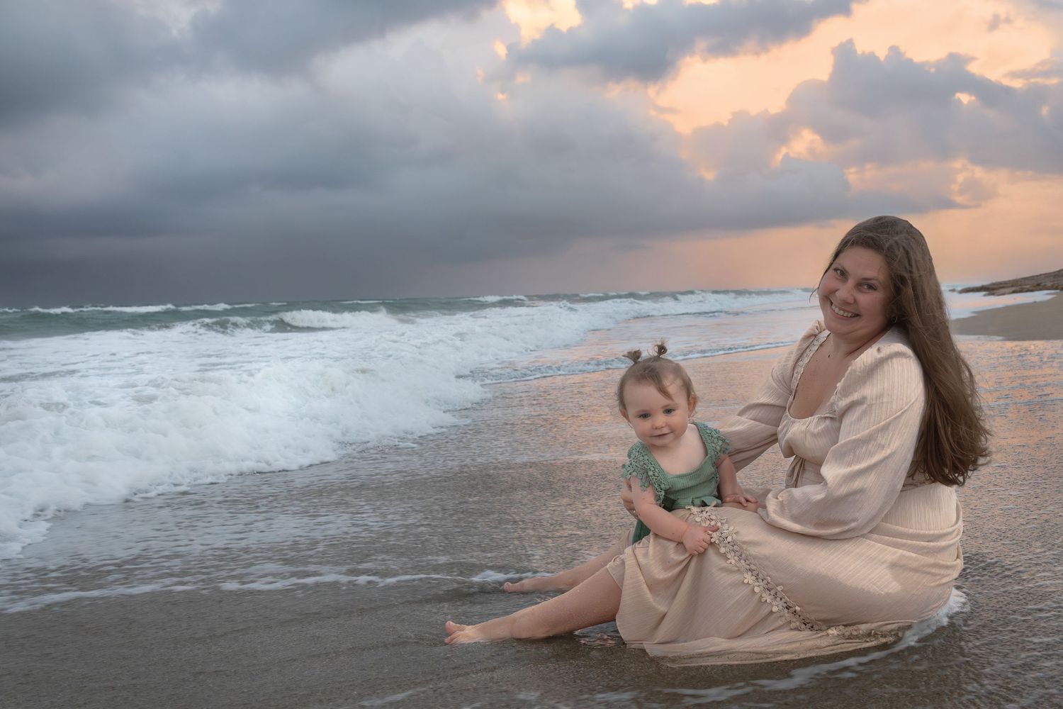 Mother and toddler sitting together on sandy beach at colorful sunset with ocean waves.