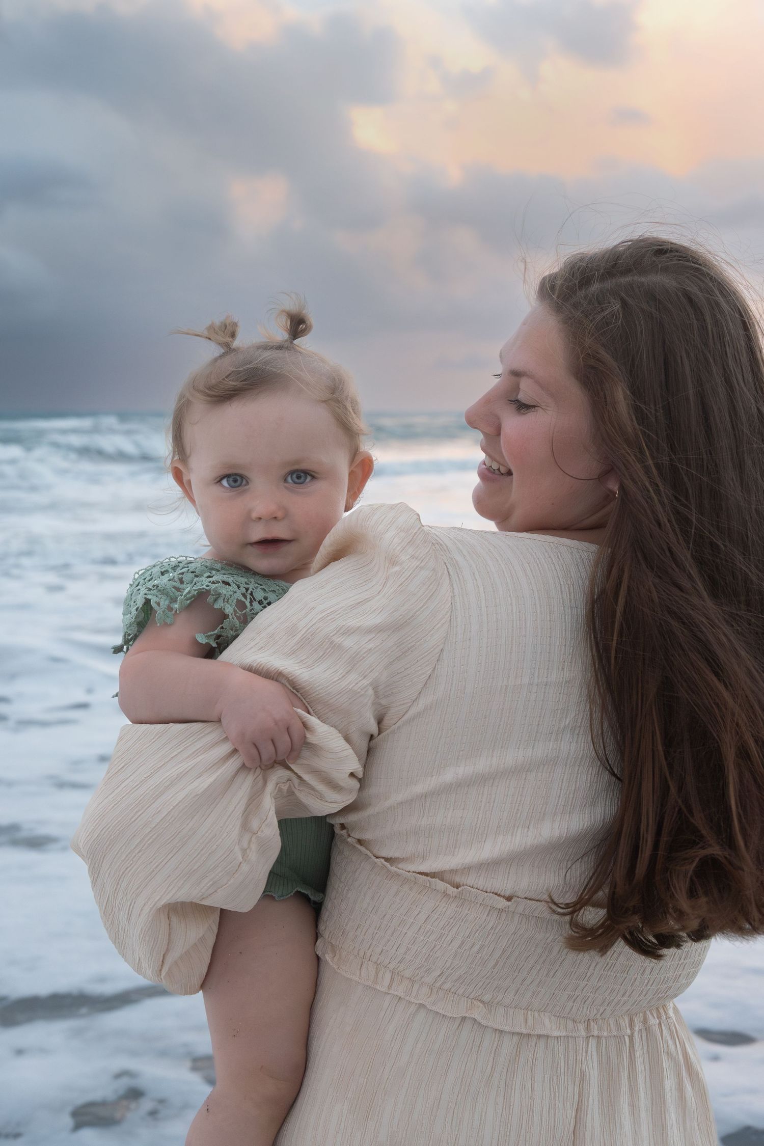 Mother holding baby with blue eyes at beach sunset, waves crashing behind them.