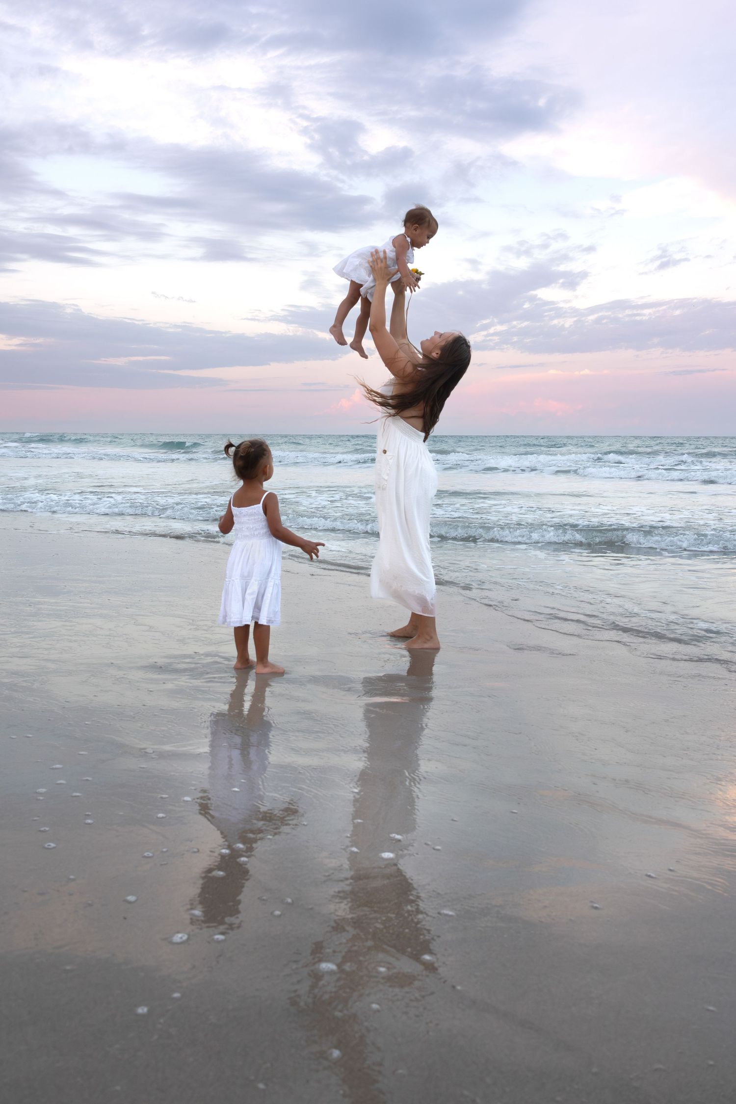 Mother lifting baby in the air on beach while toddler watches at sunset.