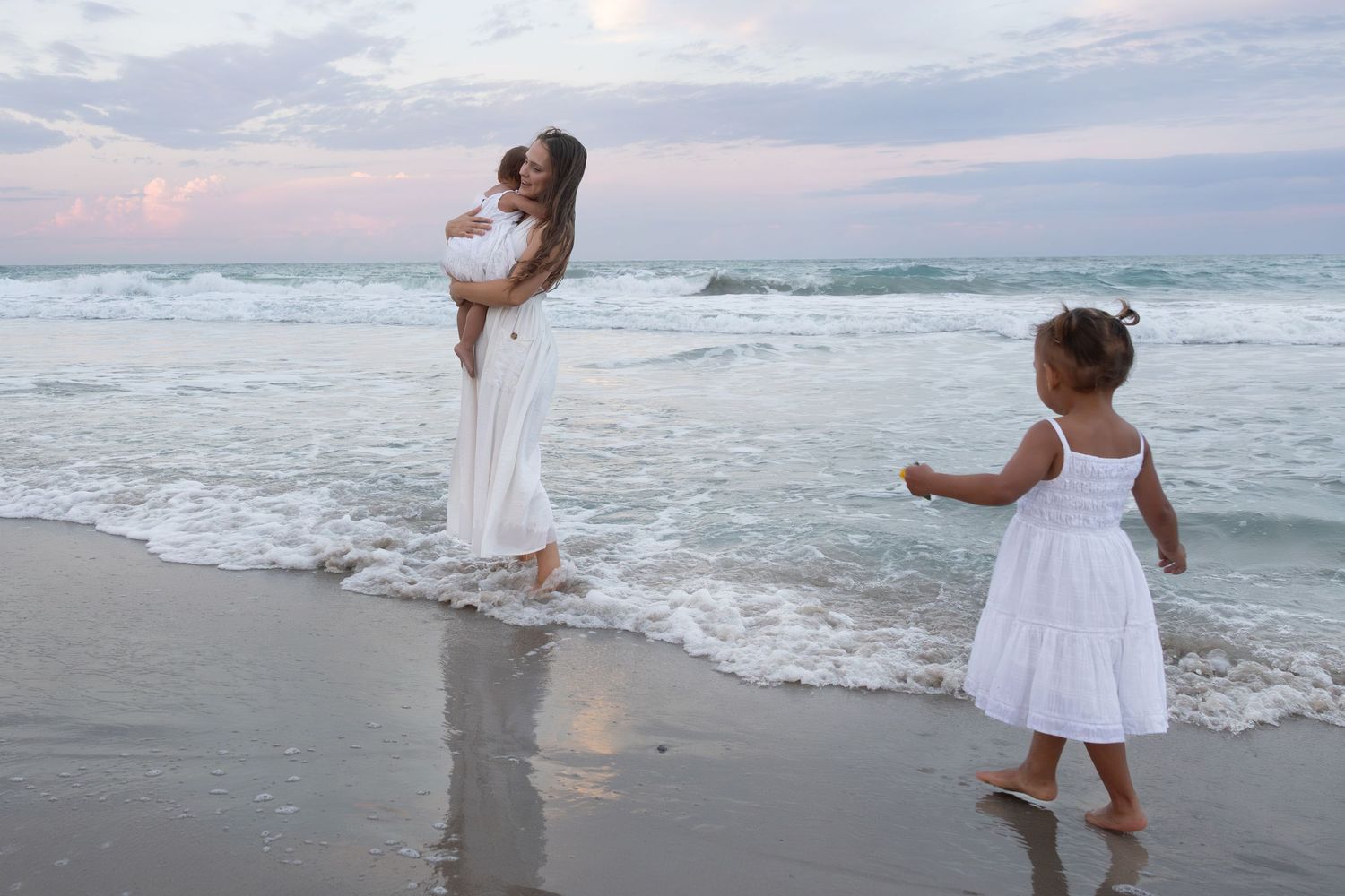 Mother holding baby while toddler dances in ocean waves at beach sunset.