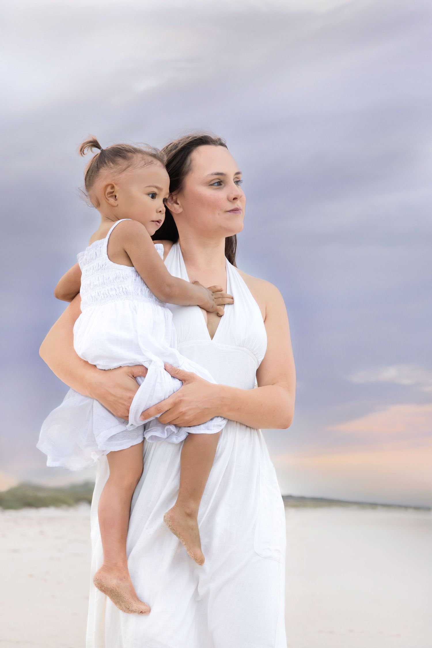 Mother in white dress holds young child on beach at sunset.