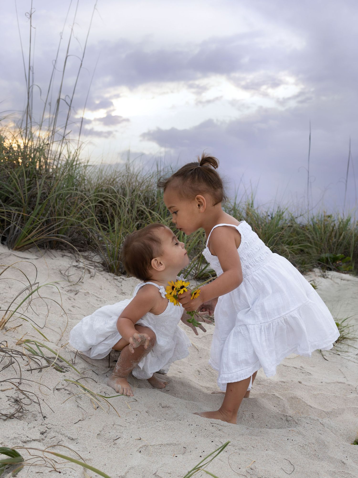 Two young girls in white dresses sharing a sweet moment on beach sand dunes.