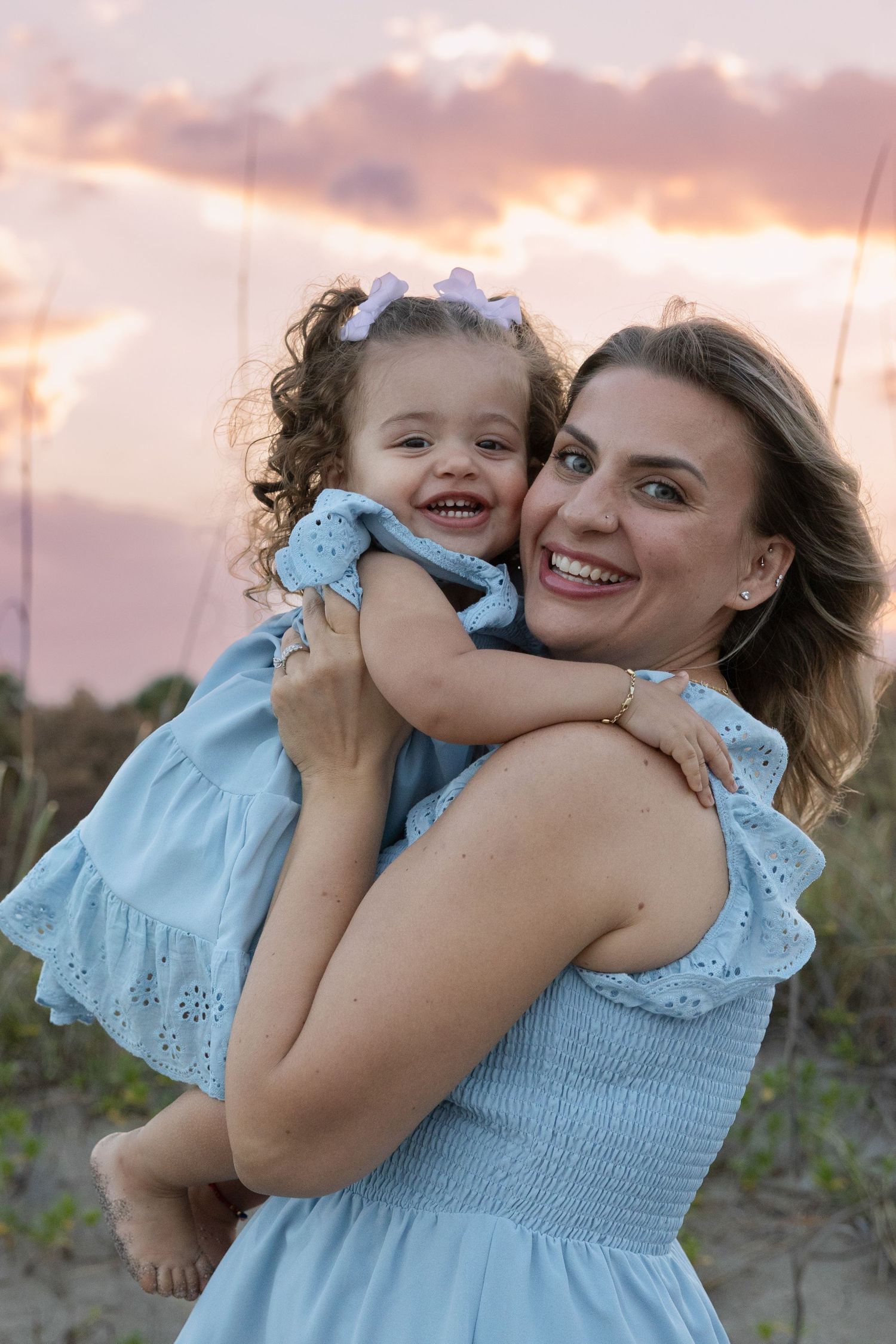 A smiling mother in a blue dress holds her laughing toddler daughter at a beach during a pink sunset.