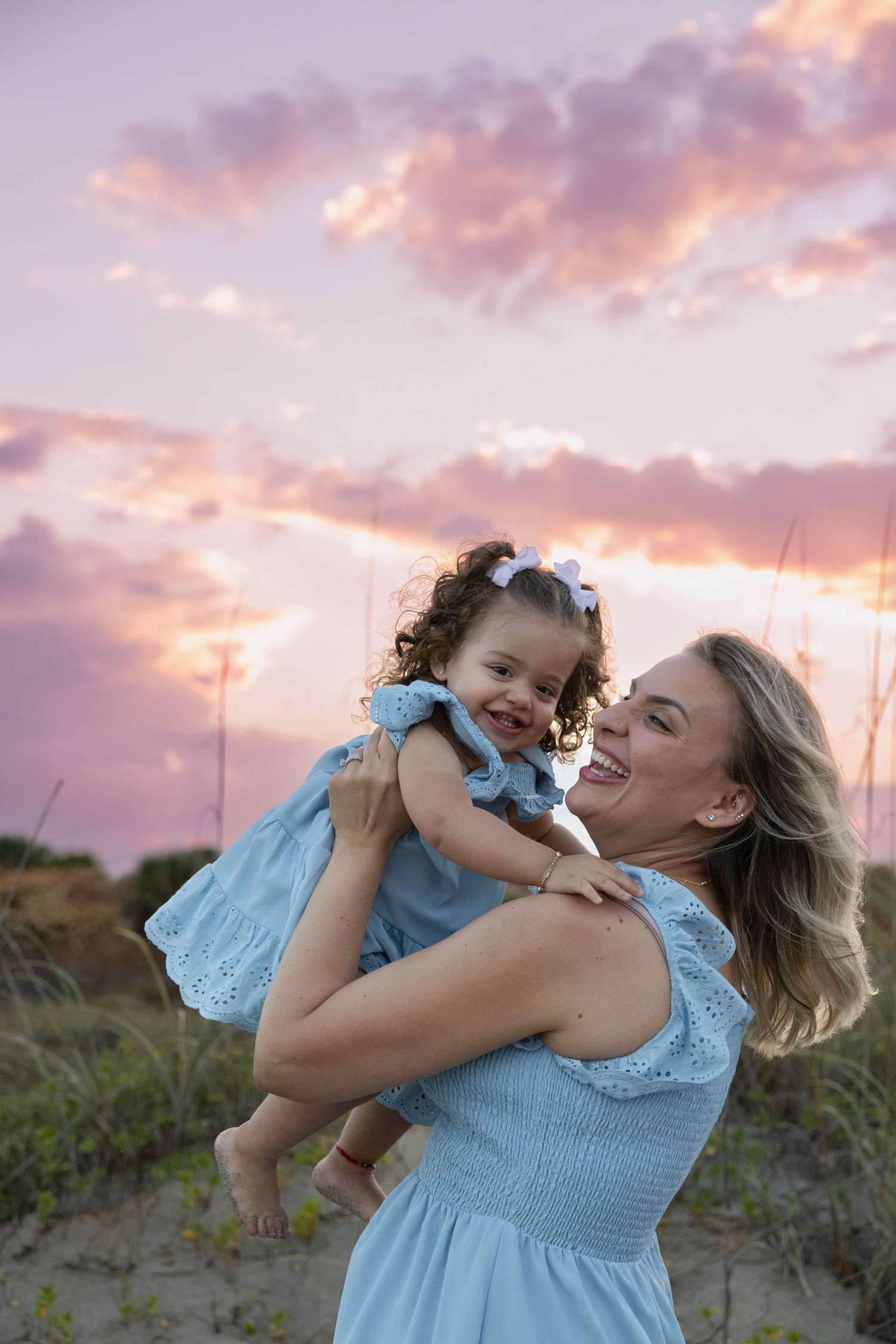 A joyful mother lifts her curly-haired daughter in matching blue dresses on a sandy beach at sunset.
