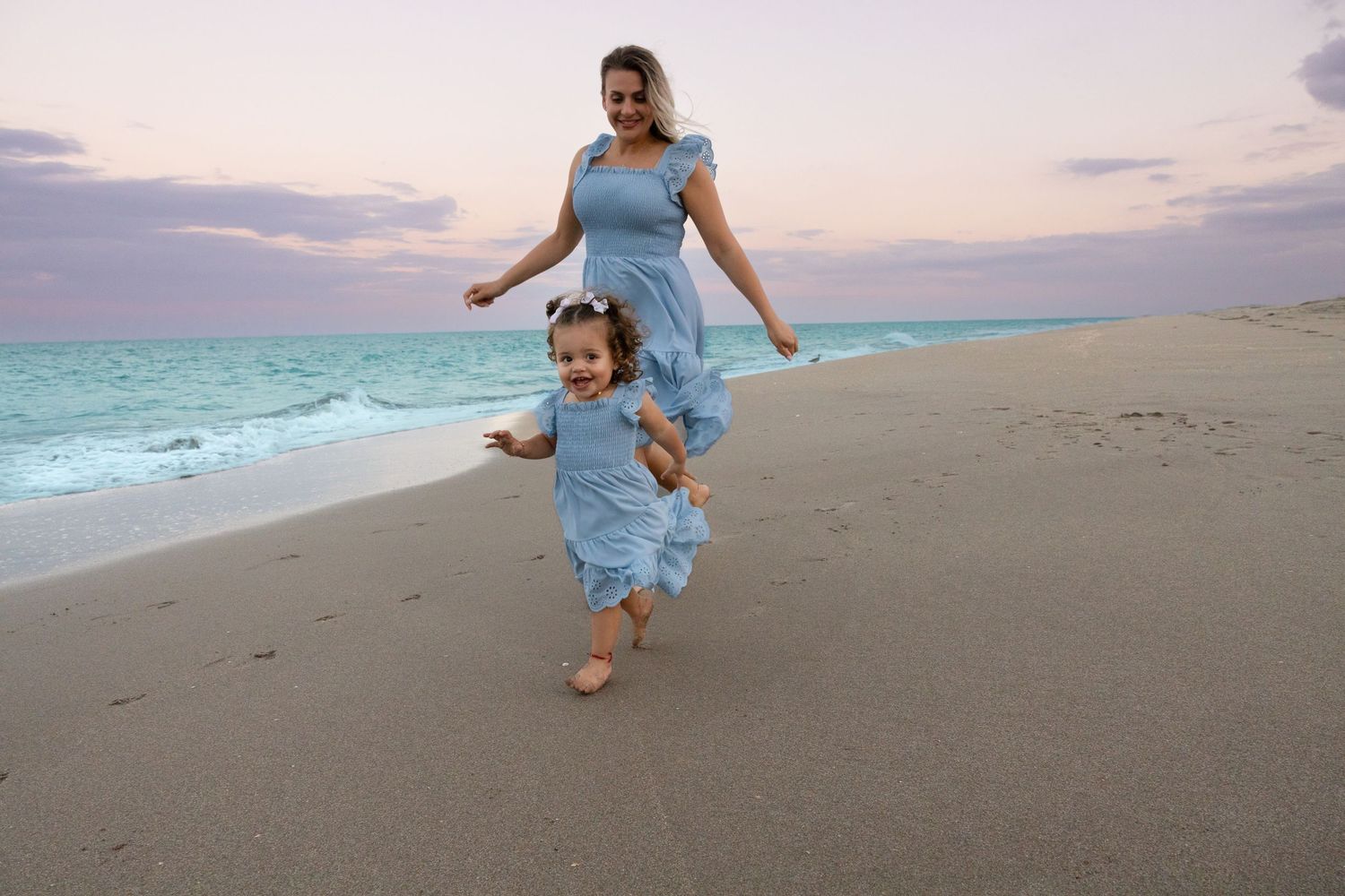 A mother and young daughter in matching blue dresses run together along a tranquil beach at sunset.