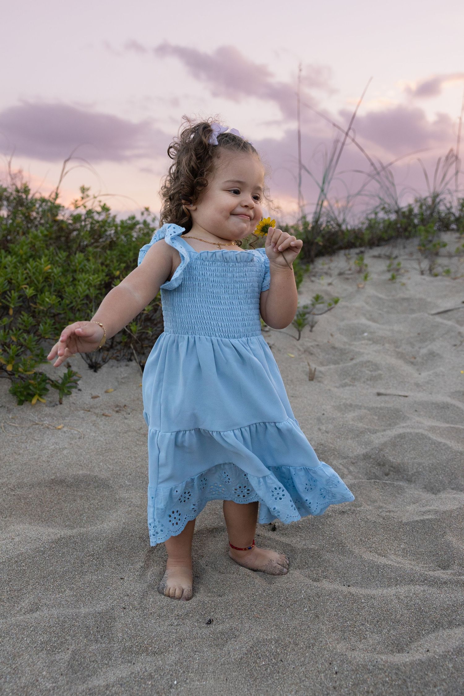 A toddler girl in a blue eyelet dress holds a yellow flower while standing on a sandy beach at dusk.