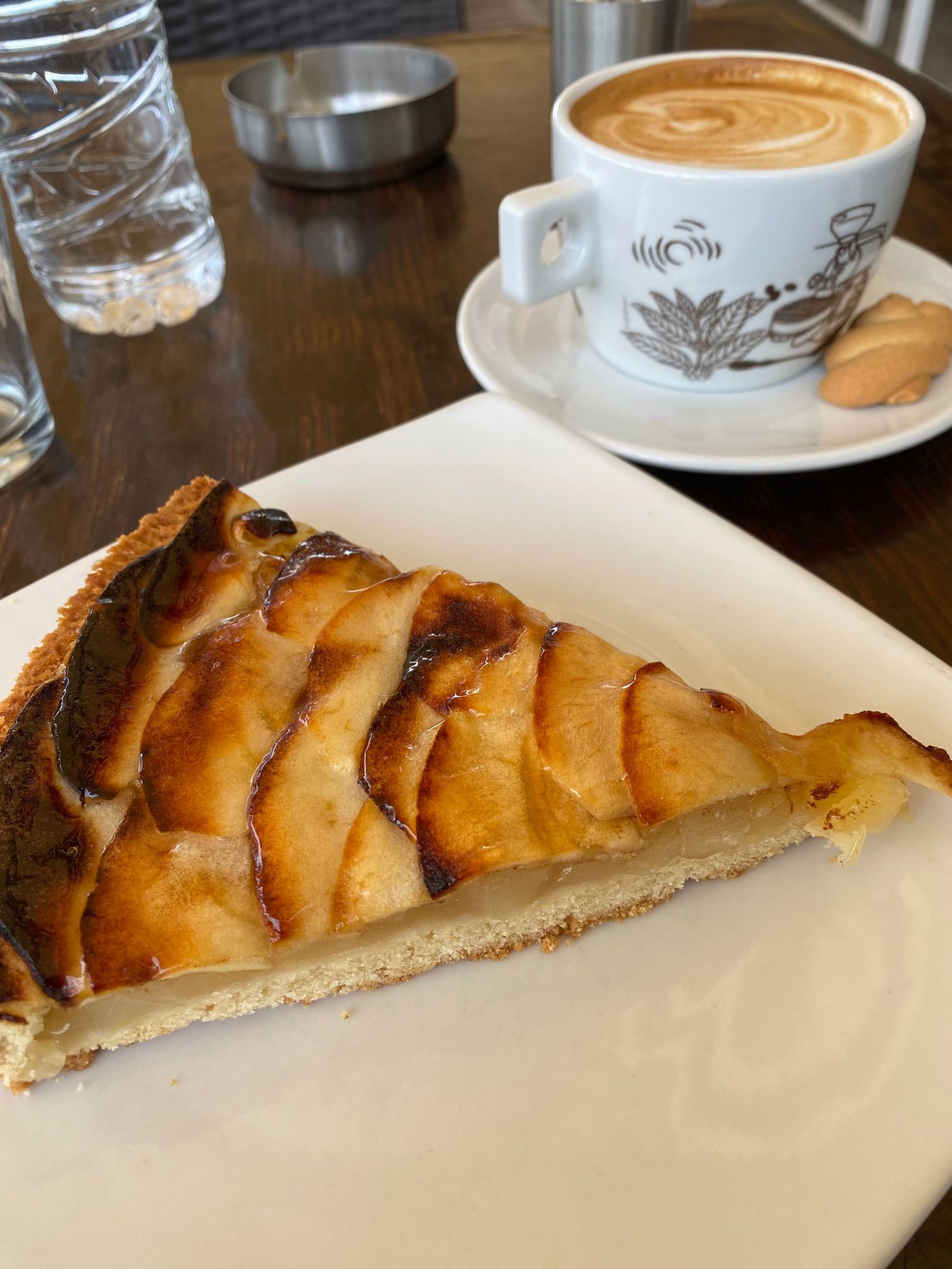 A slice of golden apple tart served on a white plate alongside a decorative coffee cup with latte art.
