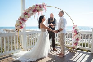 A wedding ceremony takes place on a bright oceanfront balcony decorated with pink floral arches.