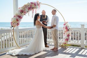 Beach wedding ceremony under a round arch decorated with pink flowers overlooking the ocean.