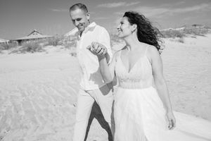 A black and white photo of a couple walking together on a beach with dunes.