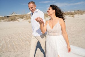 A couple walks hand in hand on a sunny beach with sand dunes in the background.