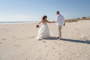 Wedding couple walks hand in hand along the shoreline during a beach photoshoot.