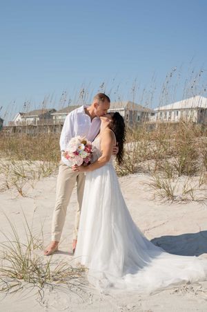 Newlyweds share an intimate moment on a sandy beach with coastal houses in the background.