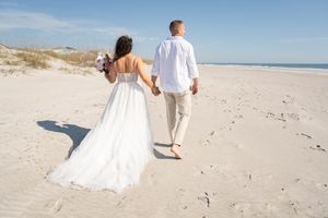 A couple walks hand in hand along a sandy beach in wedding attire.