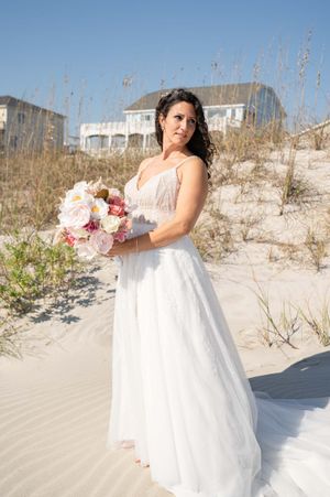 A bride in a flowing white dress poses with a pink bouquet against sandy dunes on a beach.