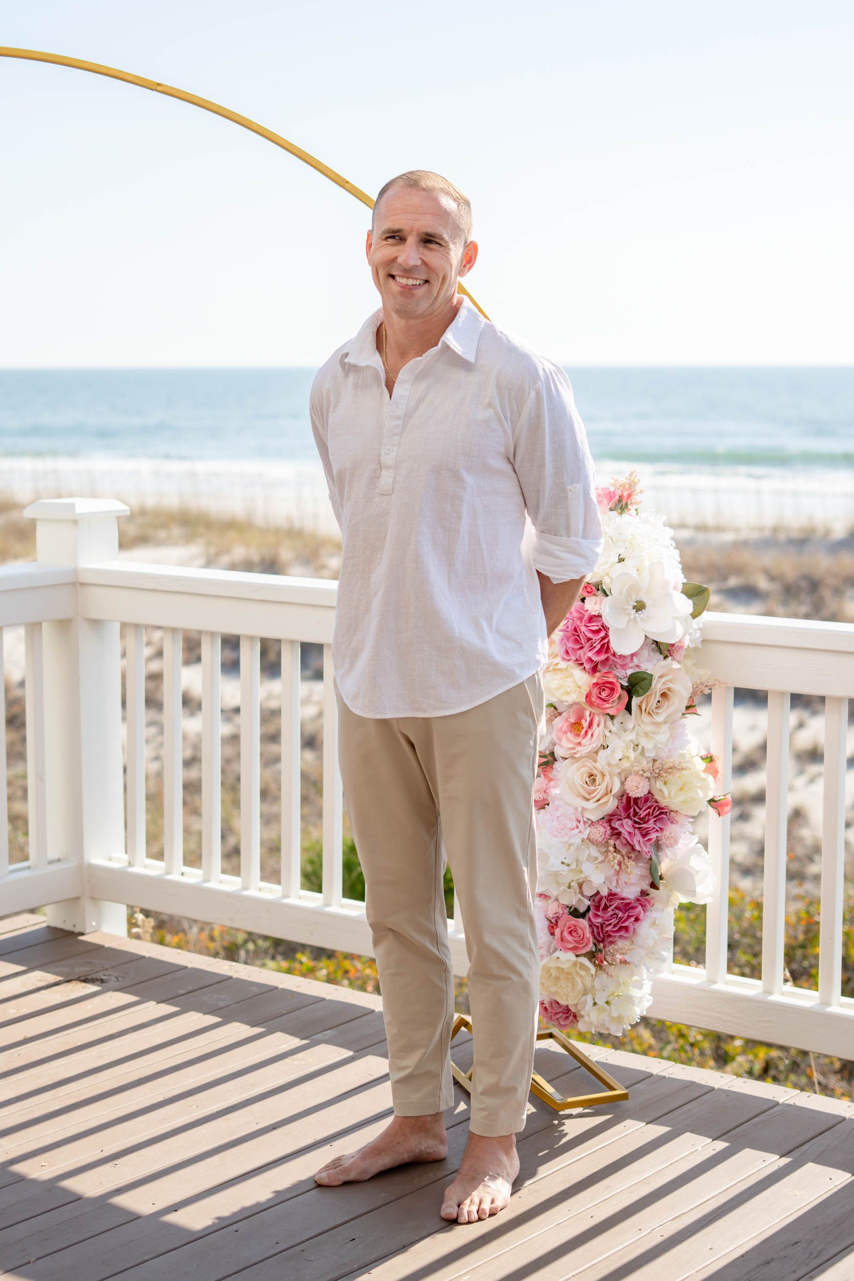 Barefoot groom in white shirt and khaki pants on beach deck with floral arrangements.