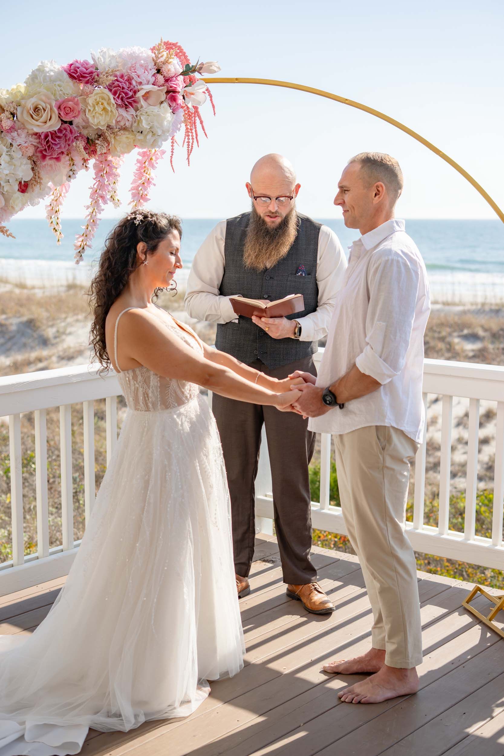 Wedding ceremony on oceanfront deck with pink floral arch and white railings.