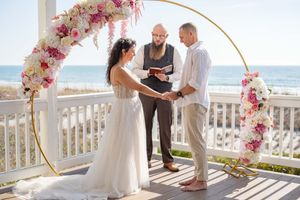 Seaside wedding ceremony with circular floral arch and white deck railing.