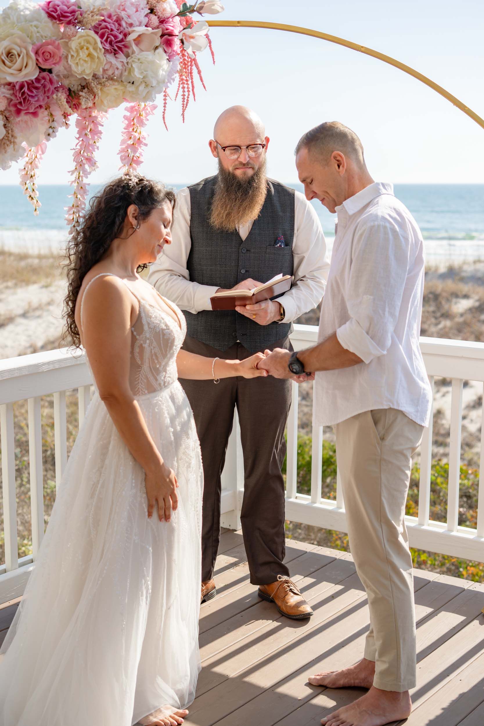 Beachfront wedding ceremony with pink and white floral arch on a wooden deck.