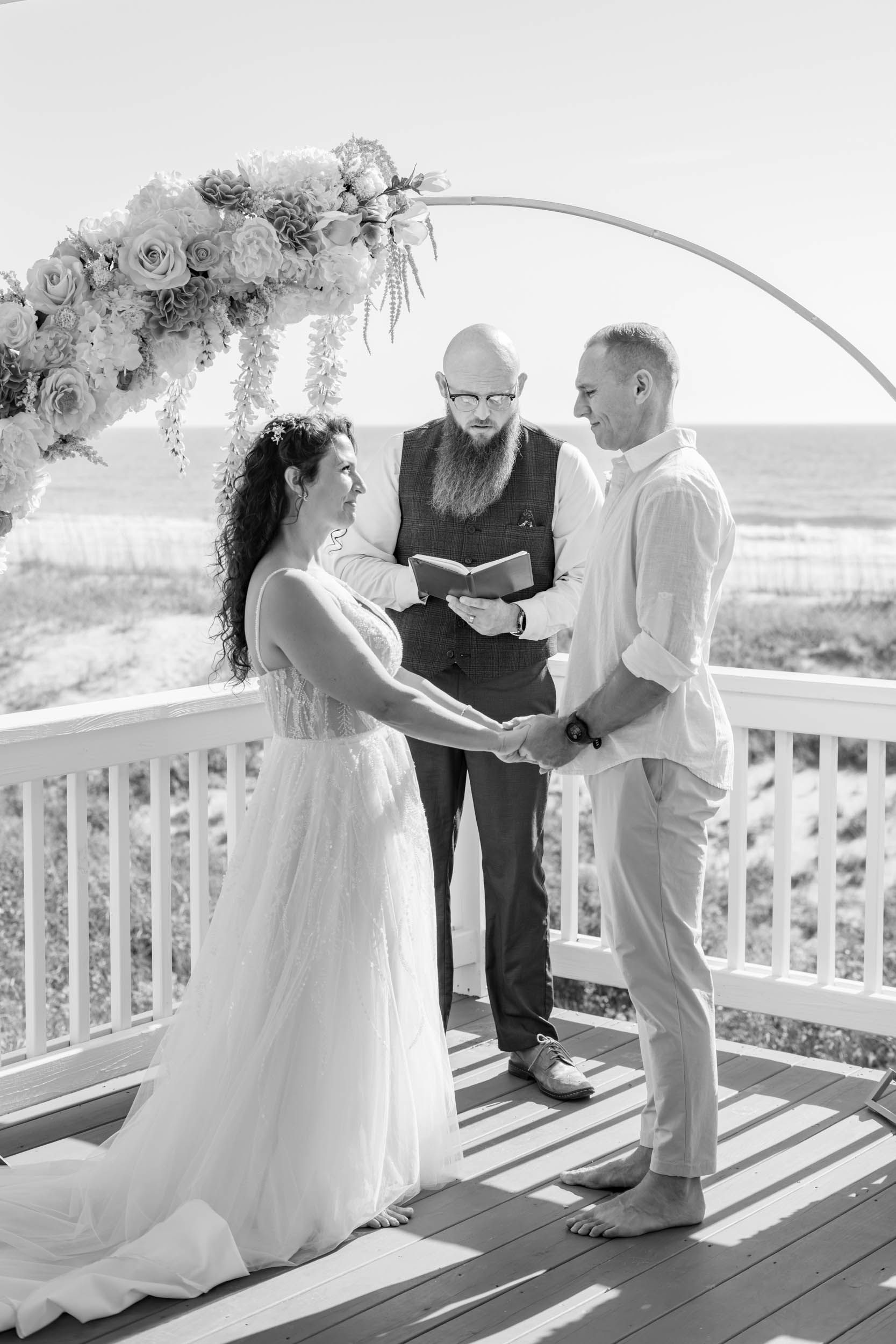 Intimate beach wedding ceremony on a white deck overlooking the ocean with floral arch decorations.