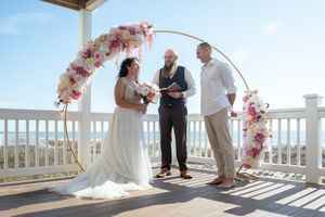 A beachside wedding ceremony unfolds under a floral arch with white railings and ocean views.