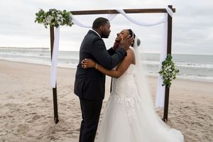 Tender moment shared between newlyweds under a wooden arch at their beach ceremony.