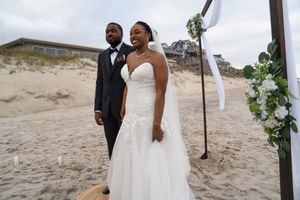 Newlyweds stand under a wooden wedding arch on a beach with sand dunes in the background.