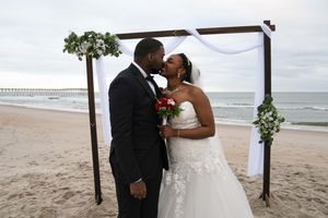 A romantic beach wedding ceremony under a wooden arch decorated with white fabric and florals by the ocean.