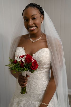 Bride in elegant white lace dress holds red roses while smiling in natural light.