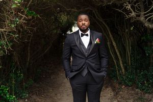Groom in black tuxedo poses against lush greenery background during outdoor wedding photoshoot.
