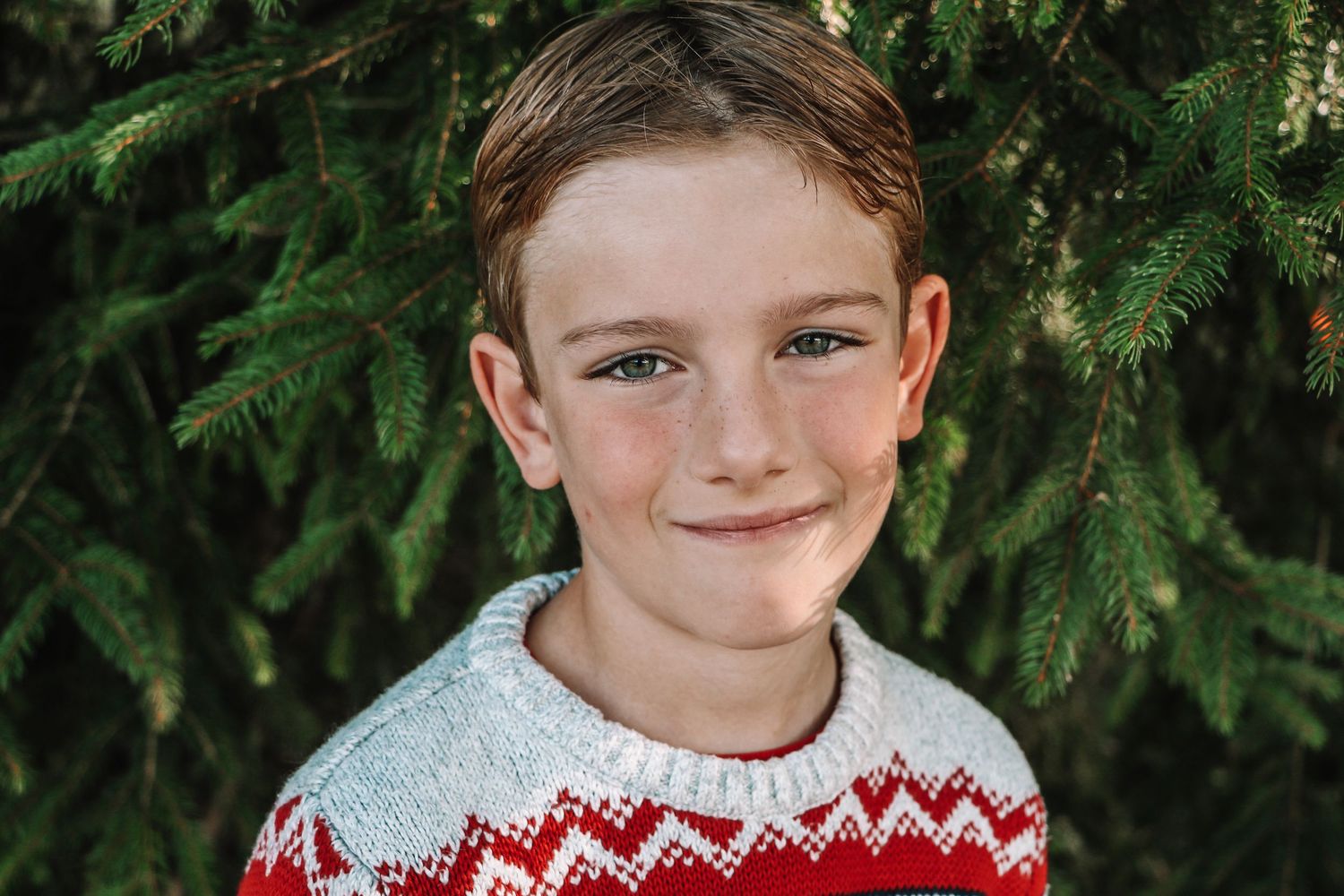 A young person in a red and white knit sweater poses outdoors against an evergreen tree background.