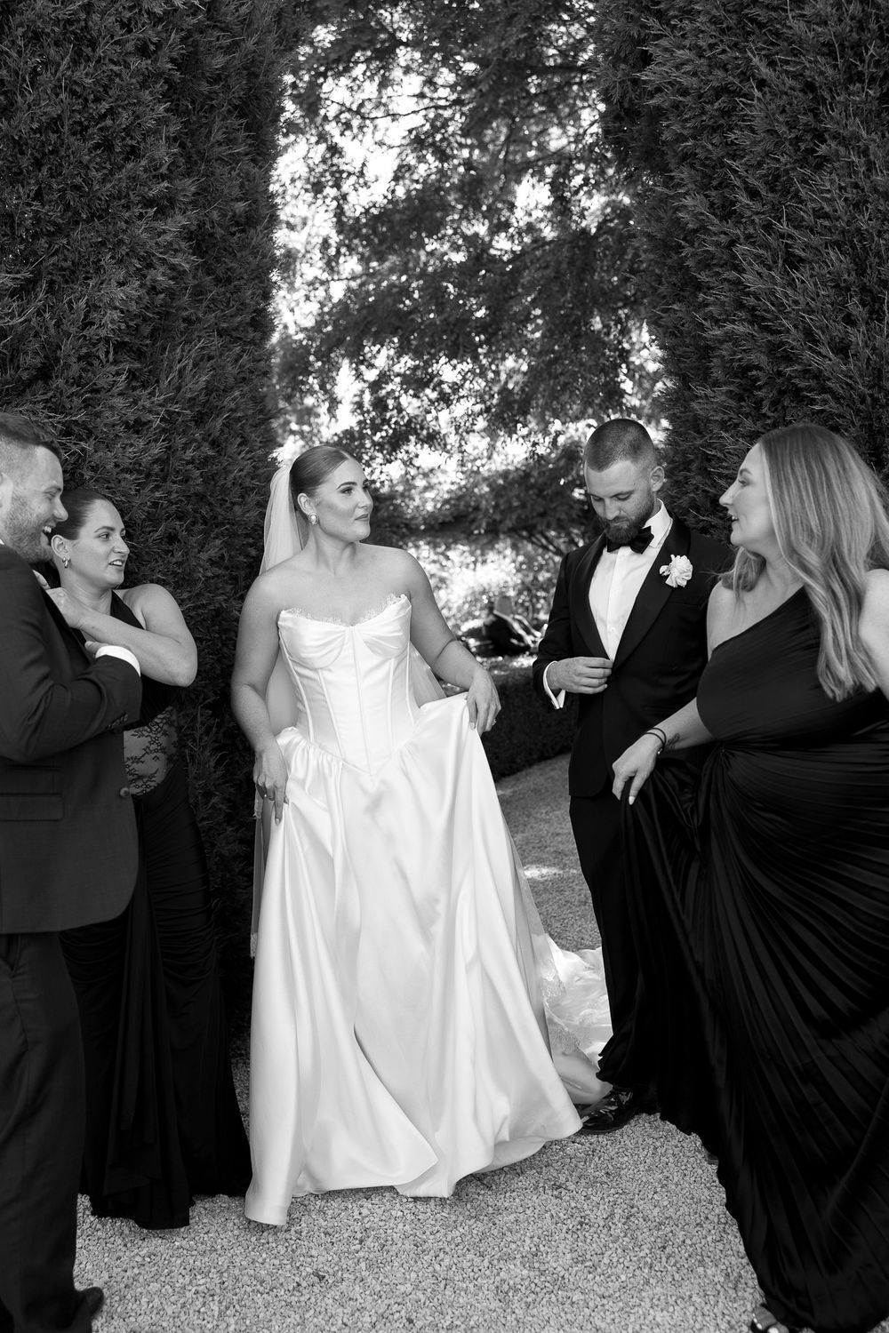 Black and white photo of a bride in a strapless gown and groom in a tuxedo surrounded by wedding guests outdoors.