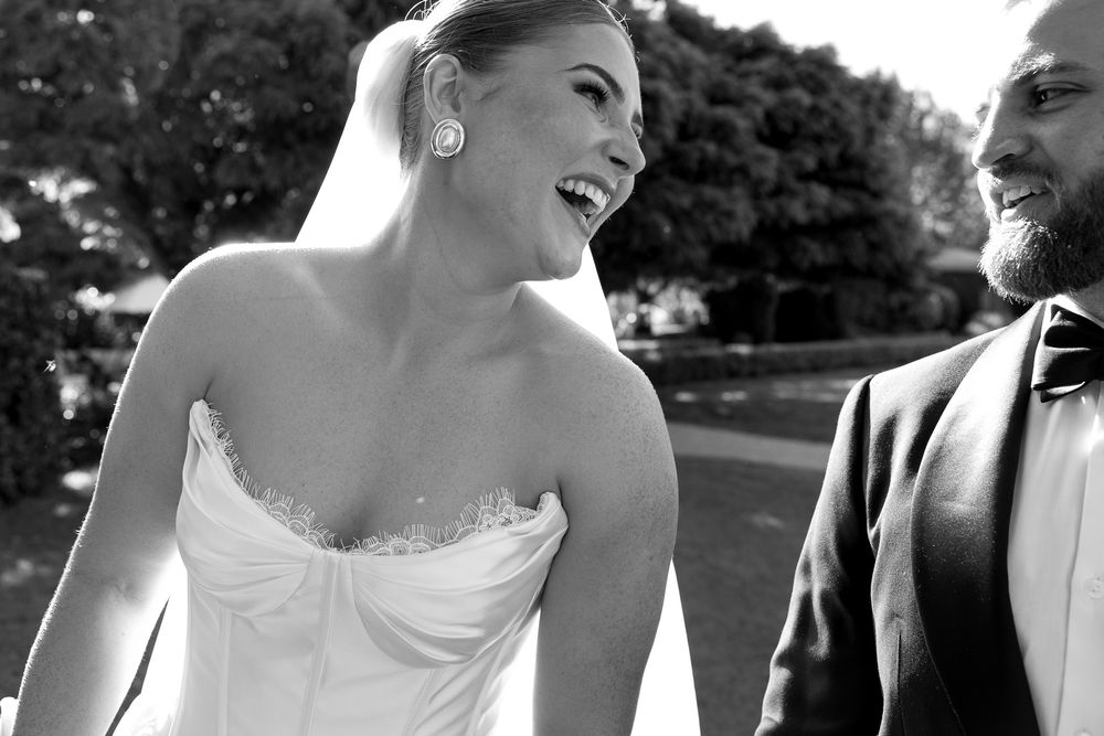 Black and white wedding photo of a laughing bride in strapless gown and groom in suit walking together outdoors.