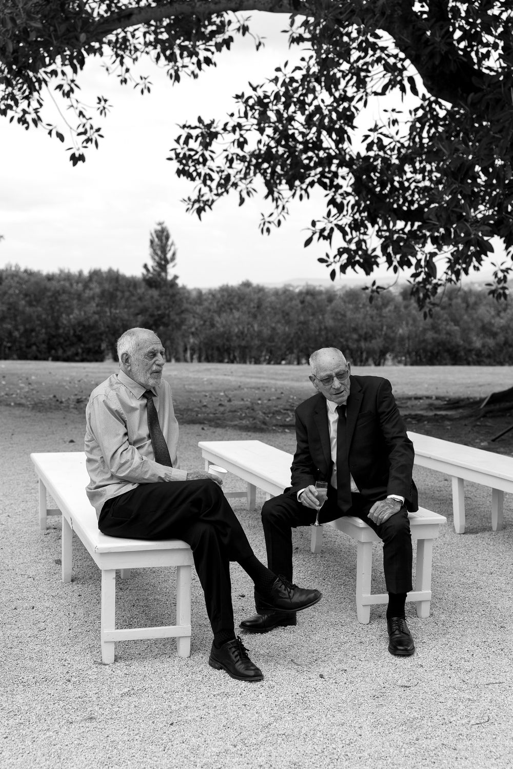 Two men sit and converse on white benches in a minimalist outdoor setting.