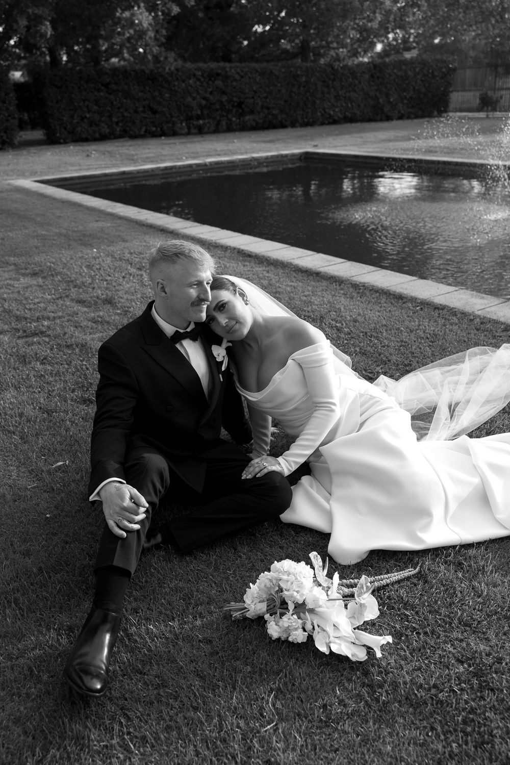 Black and white photo of bride and groom sitting on grass by a pool, bride leaning on groom in wedding attire.