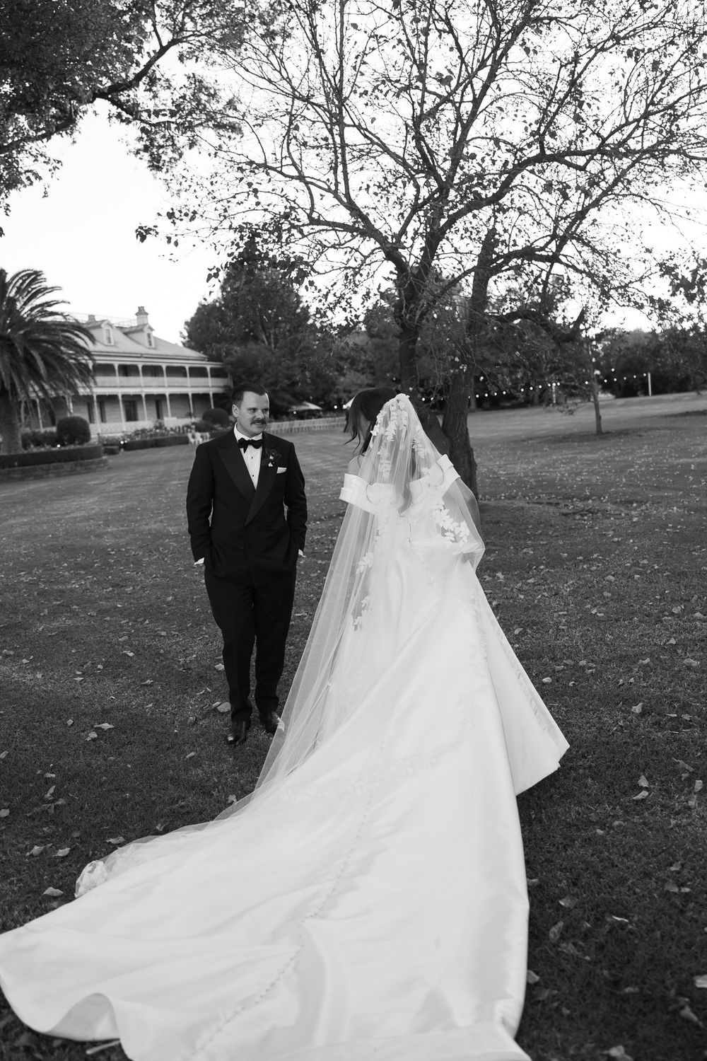 Black and white photo of bride and groom walking outdoors on wedding day, bride wearing long veil and gown.