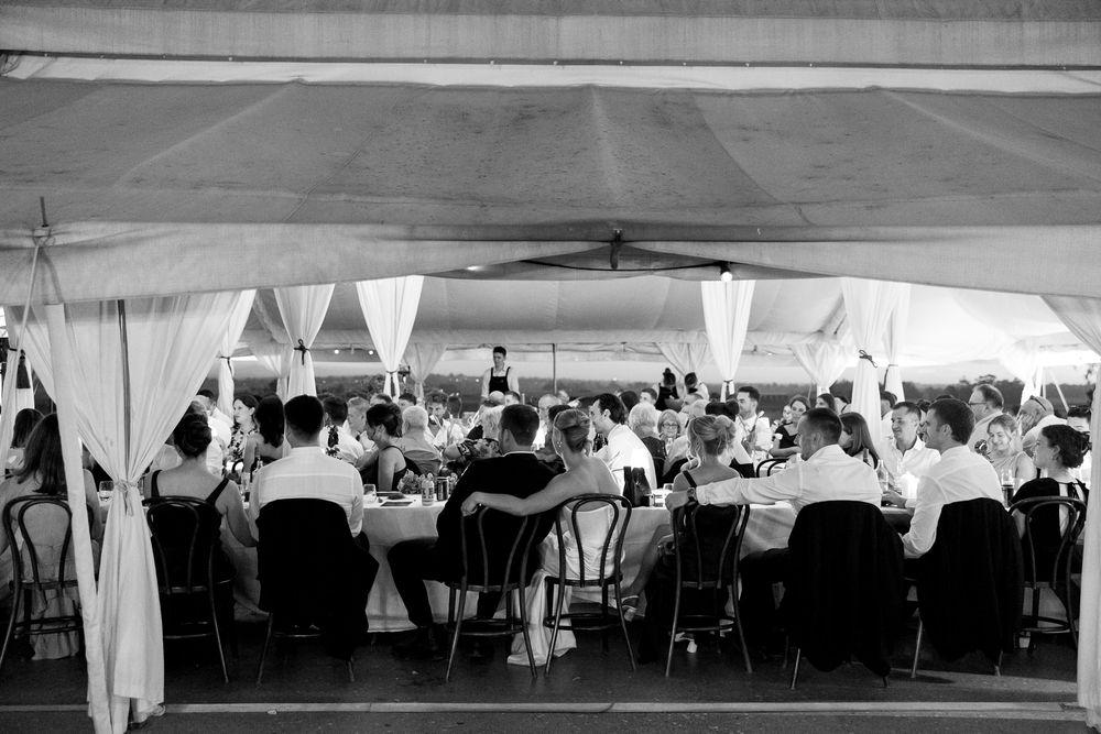 Black and white photo of elegantly dressed guests dining under a draped tent at a formal outdoor wedding reception.