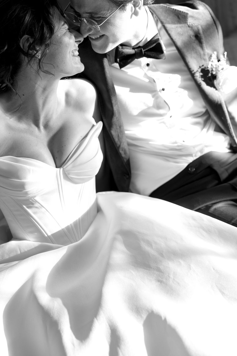 Black and white photo of a smiling bride in a white gown and groom in a bow tie sharing a joyful moment.