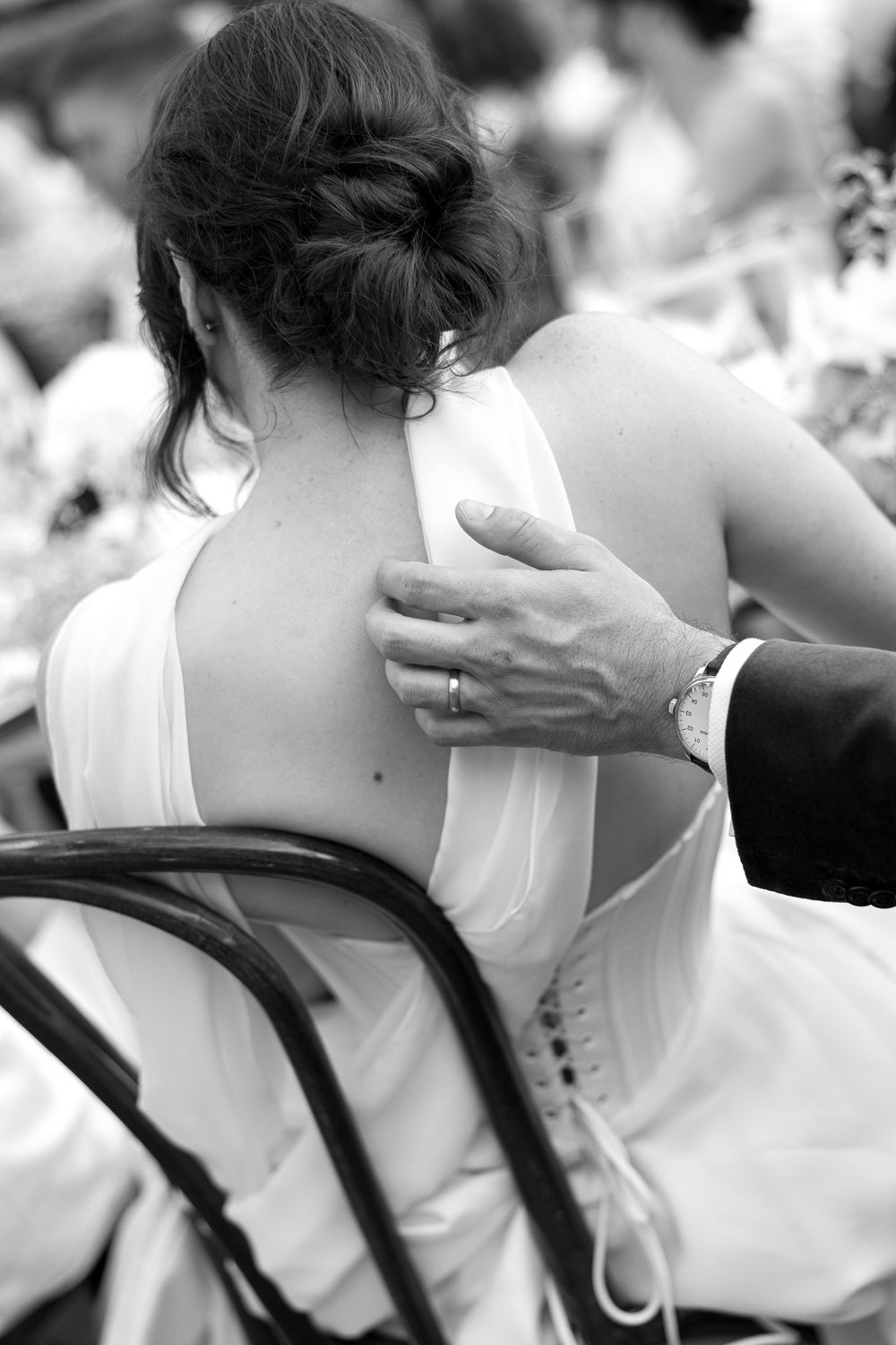 Black and white photo of a bride in a backless white gown with groom's hand on her shoulder at a wedding reception.