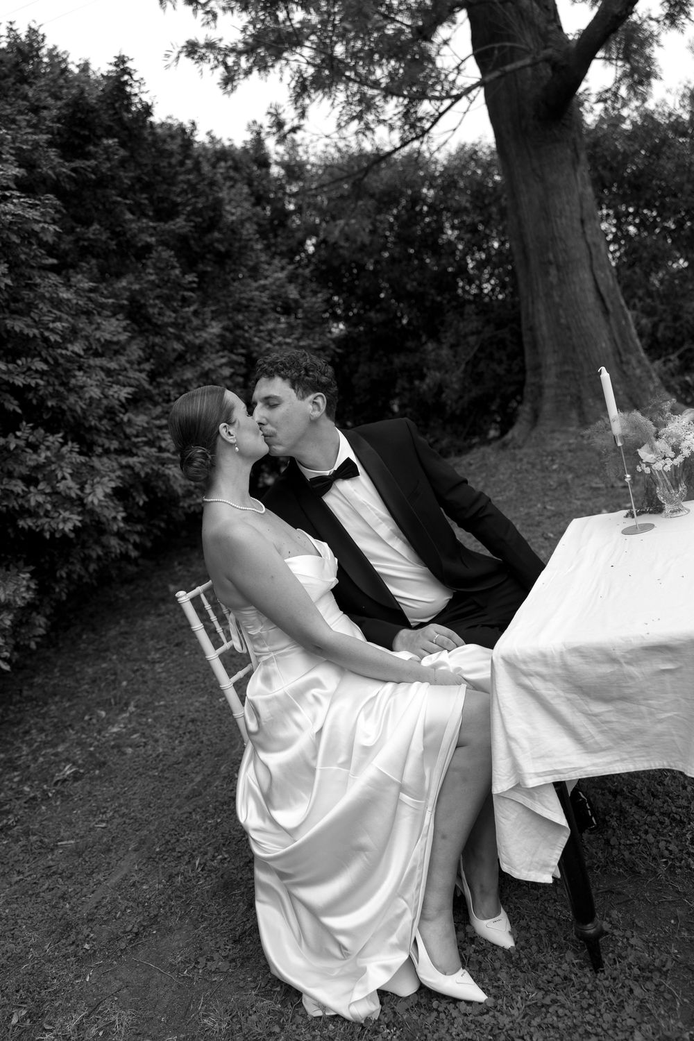 A romantic black and white photo series showing a couple in white attire sitting on a tree branch in a park setting.